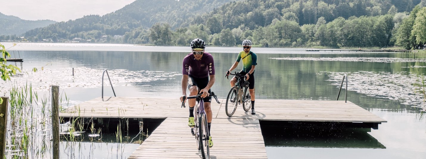 Gravelbike Urlaub in Kärnten © Region Wörthersee Rosental Tourismus GmbH - Chris Perkles Zwei Radfahrer fahren auf einem Steg über einen ruhigen See mit Bergen im Hintergrund