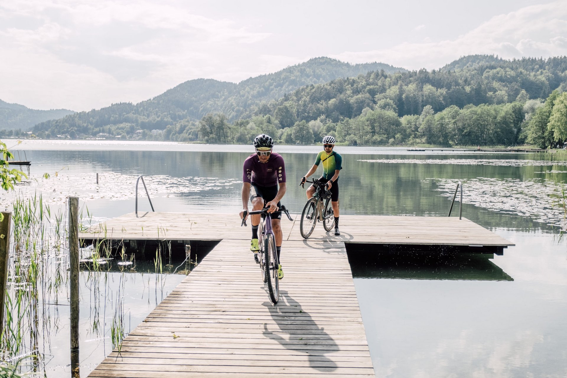 Two cyclists riding on a wooden dock over a calm lake with mountains behind