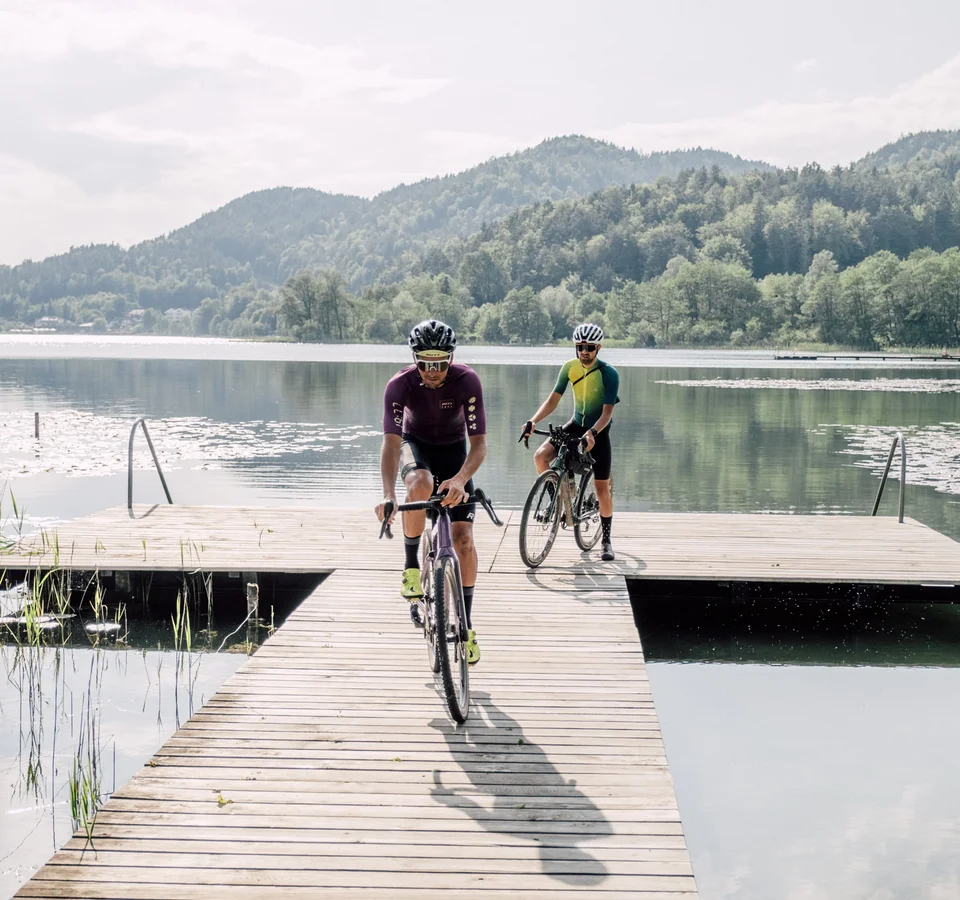 Zwei Radfahrer fahren auf einem Steg über einen ruhigen See mit Bergen im Hintergrund