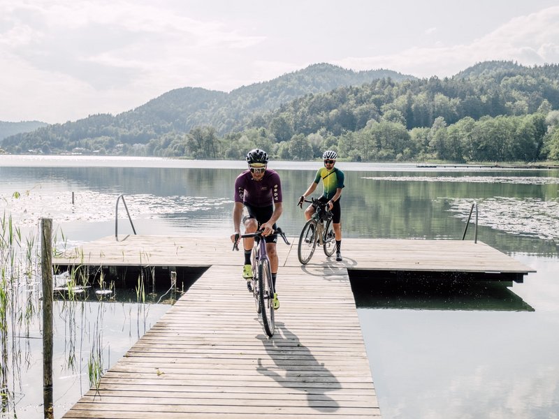 Zwei Radfahrer fahren auf einem Steg über einen ruhigen See mit Bergen im Hintergrund