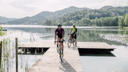 Two cyclists riding on a wooden dock over a calm lake with mountains behind
