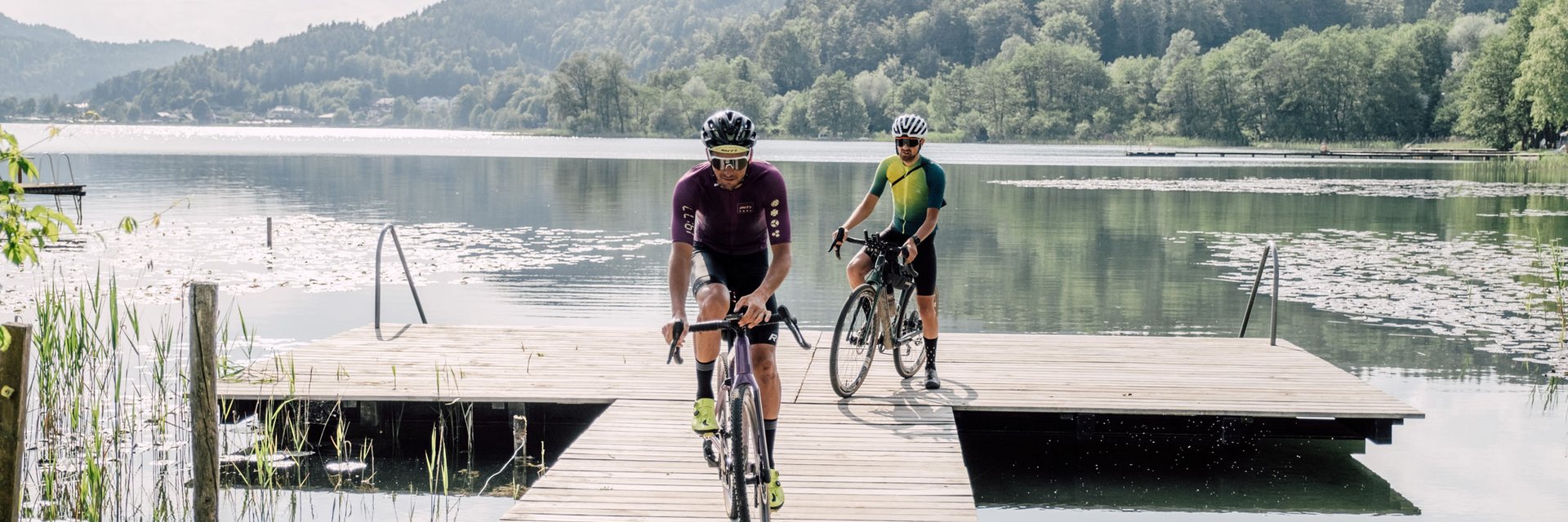 Two cyclists riding on a wooden dock over a calm lake with mountains behind