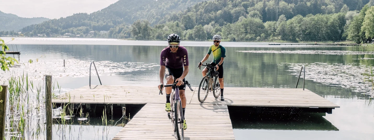 Two cyclists riding on a wooden dock over a calm lake with mountains behind