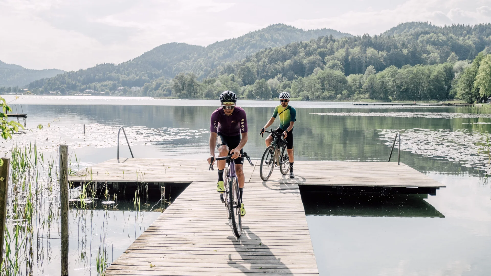 Two cyclists riding on a wooden dock over a calm lake with mountains behind