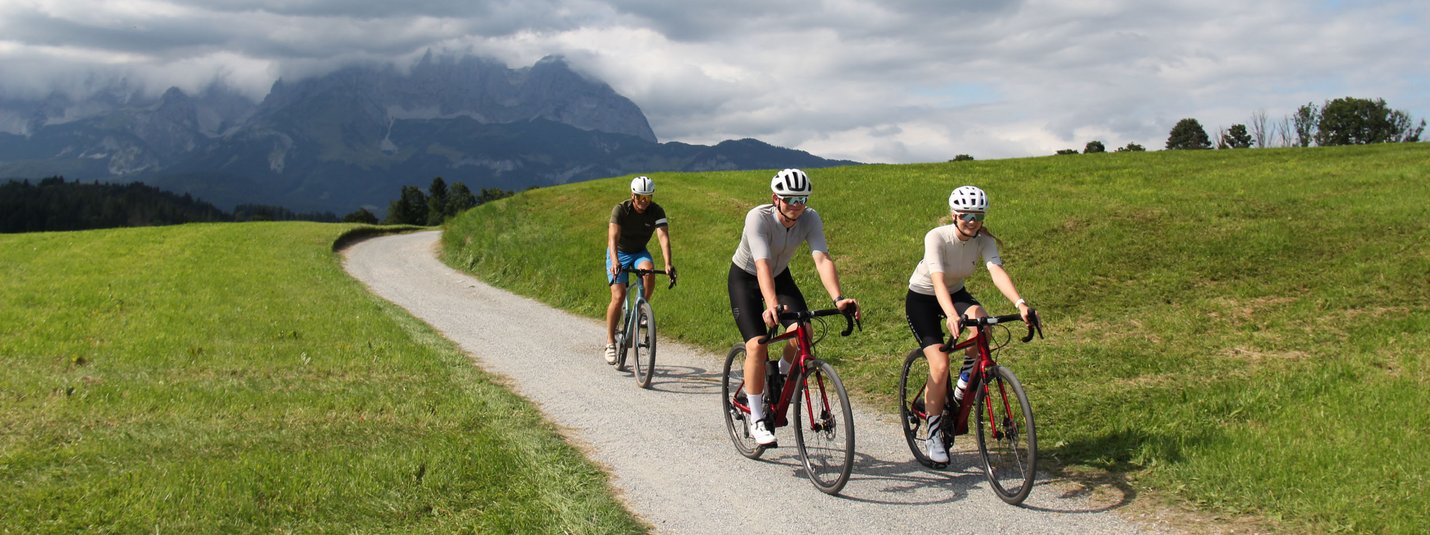 Drei Radfahrer auf Landstraße mit Bergen und Wolken im Hintergrund