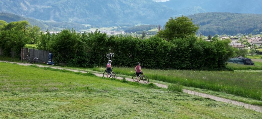 Two cyclists on a path through green fields with mountains in the background