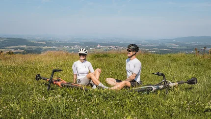 Two cyclists sitting on grass on a hill overlooking a landscape.