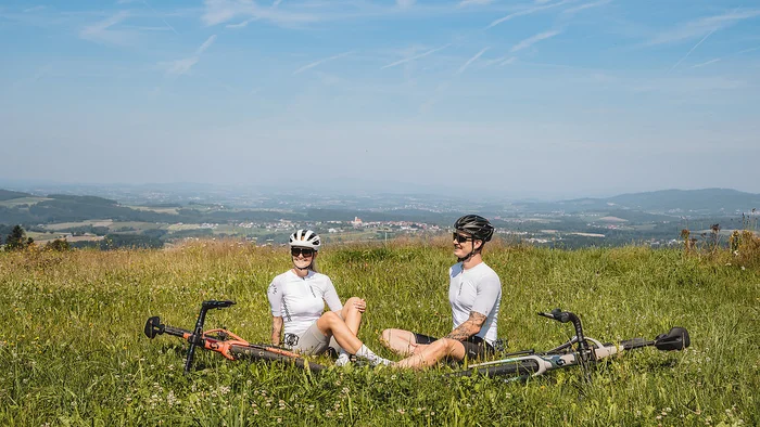 Zwei Radfahrer sitzen im Gras auf einem Hügel mit Blick auf eine Landschaft.