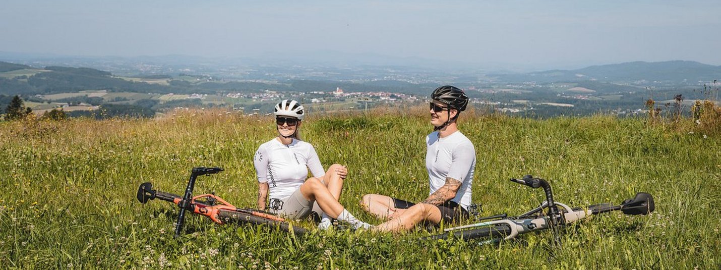 Gravelbike Urlaub in Bayern © Stefan J. Wolf Photography Zwei Radfahrer sitzen im Gras auf einem Hügel mit Blick auf eine Landschaft.