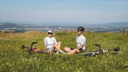Zwei Radfahrer sitzen im Gras auf einem Hügel mit Blick auf eine Landschaft.