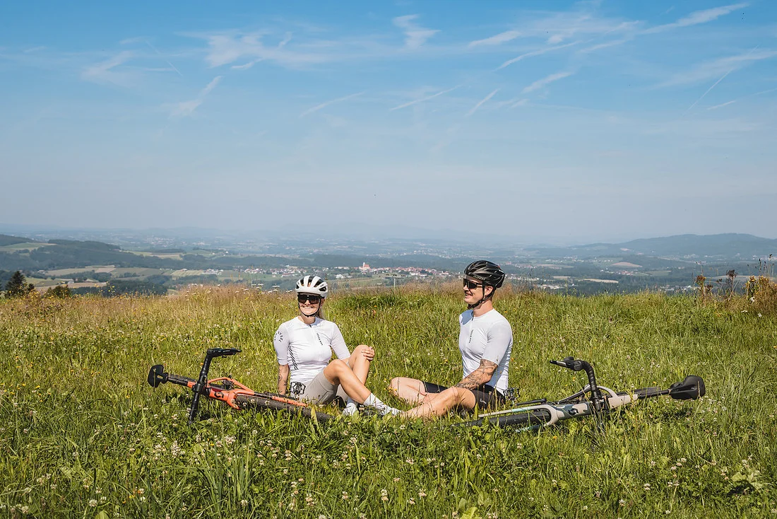 Gravelbiken im Bayerischen Wald © Stefan J. Wolf Photography Zwei Radfahrer sitzen im Gras auf einem Hügel mit Blick auf eine Landschaft.