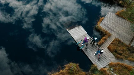 Three cyclists on a dock by a lake with clouds reflected in the water