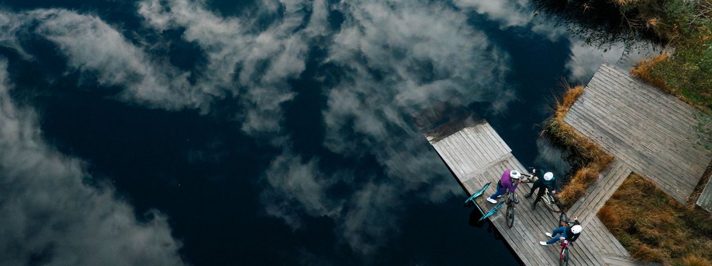 Three cyclists on a dock by a lake with clouds reflected in the water