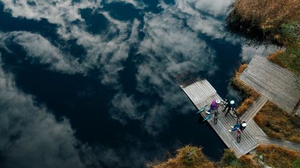 Drei Radfahrer auf einem Steg am See mit Wolkenreflektion im Wasser