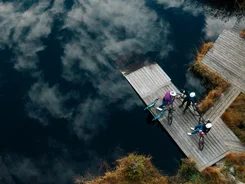 Three cyclists on a dock by a lake with clouds reflected in the water