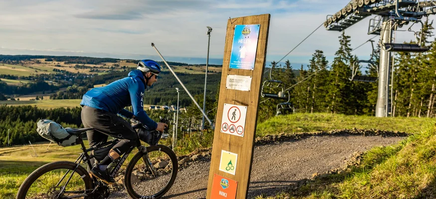 Fahrradfahrer auf Waldweg neben einem Schlepplift und Wegweiser in Alpenlandschaft