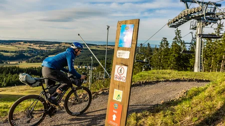 Fahrradfahrer auf Waldweg neben einem Schlepplift und Wegweiser in Alpenlandschaft