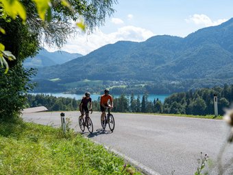 Two cyclists on a country road with mountains and lake in the background