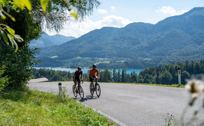 Gravelbike Urlaub in der Fuschlseeregion © Christina Neubauer Zwei Radfahrer auf einer Landstraße mit Berg- und Seehintergrund