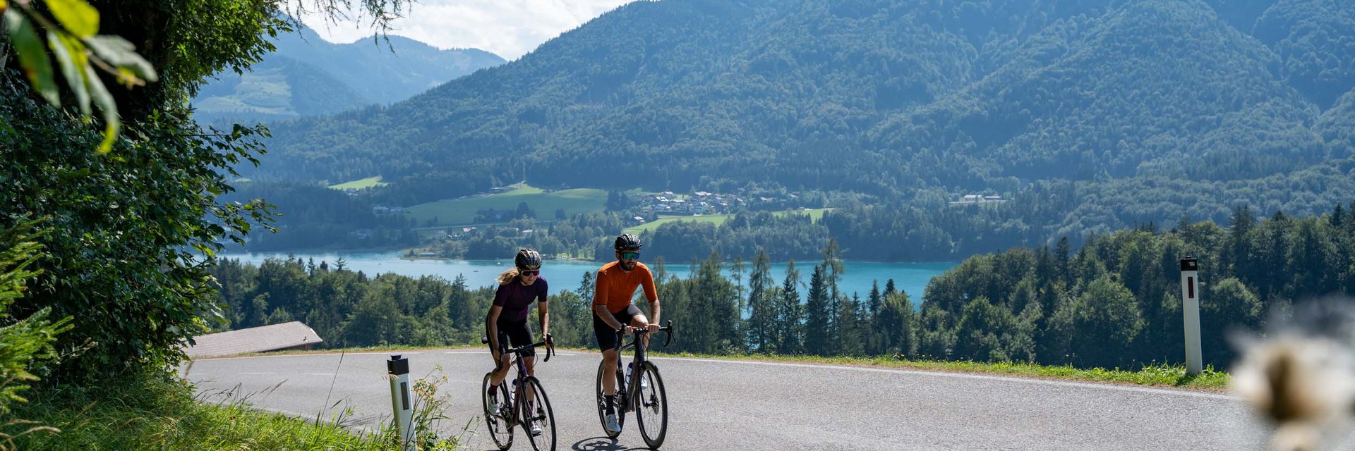 Zwei Radfahrer auf einer Landstraße mit Berg- und Seehintergrund