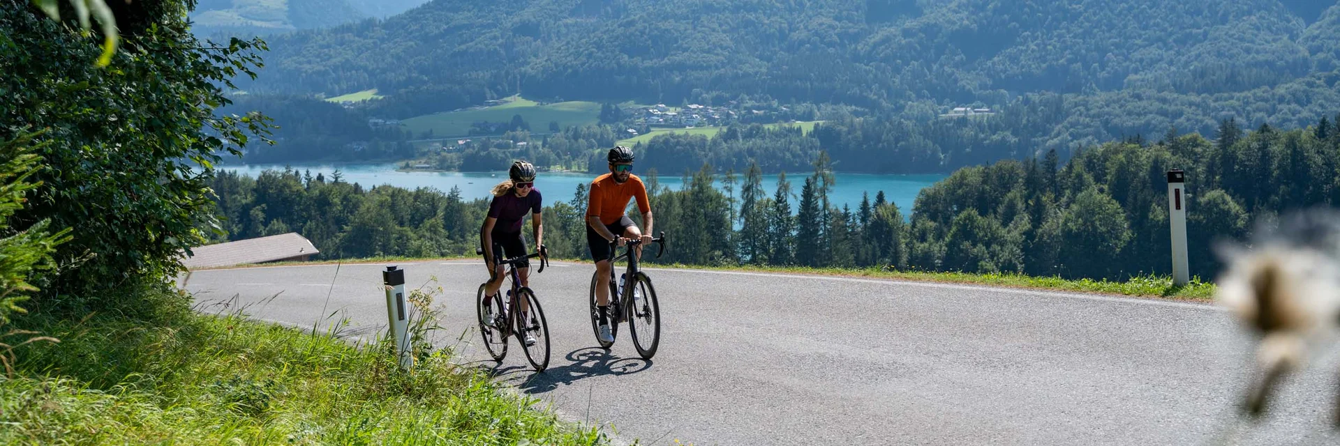 Two cyclists on a country road with mountains and lake in the background
