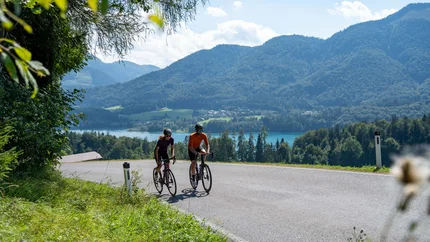 Two cyclists on a country road with mountains and lake in the background