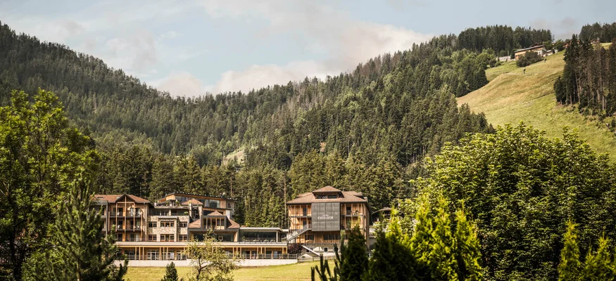 Herbst auf Schotter im Excelsior Dolomites Life Resort****s © Hannes Niederkofler Hotelgebäude eingebettet in grüne Berge und Wald unter blauem Himmel