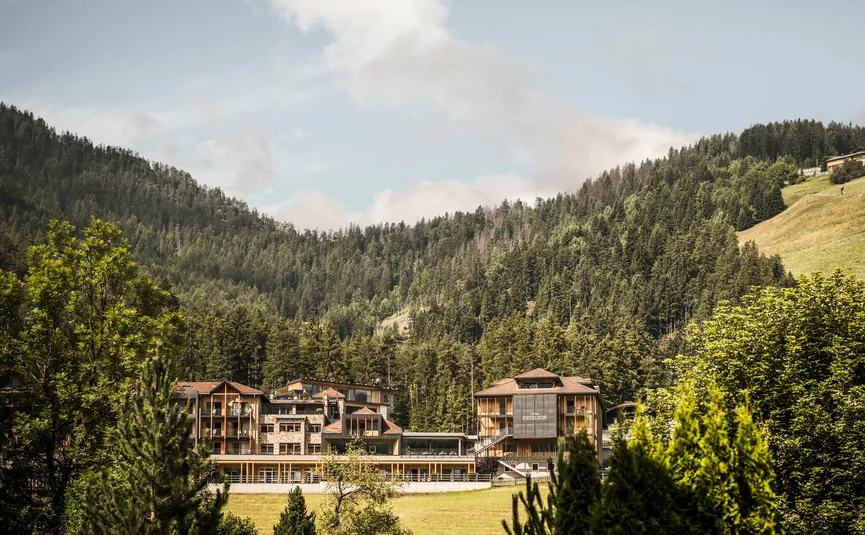 Excelsior Dolomites Life Resort ****S © Hannes Niederkofler Hotelgebäude eingebettet in grüne Berge und Wald unter blauem Himmel