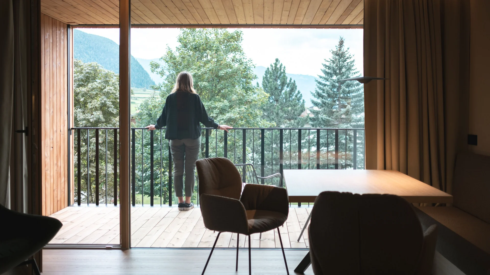 Neue Gravelbike Holidays Hotels 2026 © Lunah Photography - Lukas Haberer & Hannah Beikircher Person blickt vom Balkon eines Holzhauses auf grüne Bäume und Berge