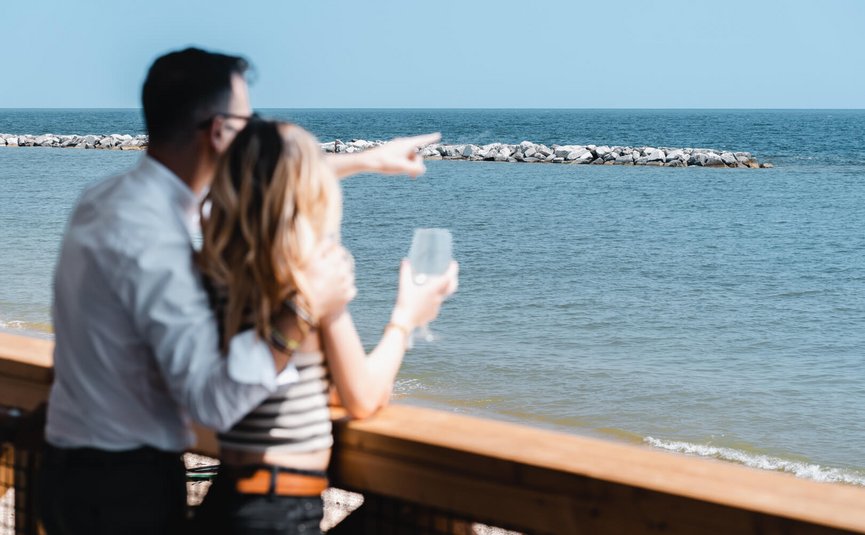Couple on terrace by sea, man pointing at water, woman holding glass