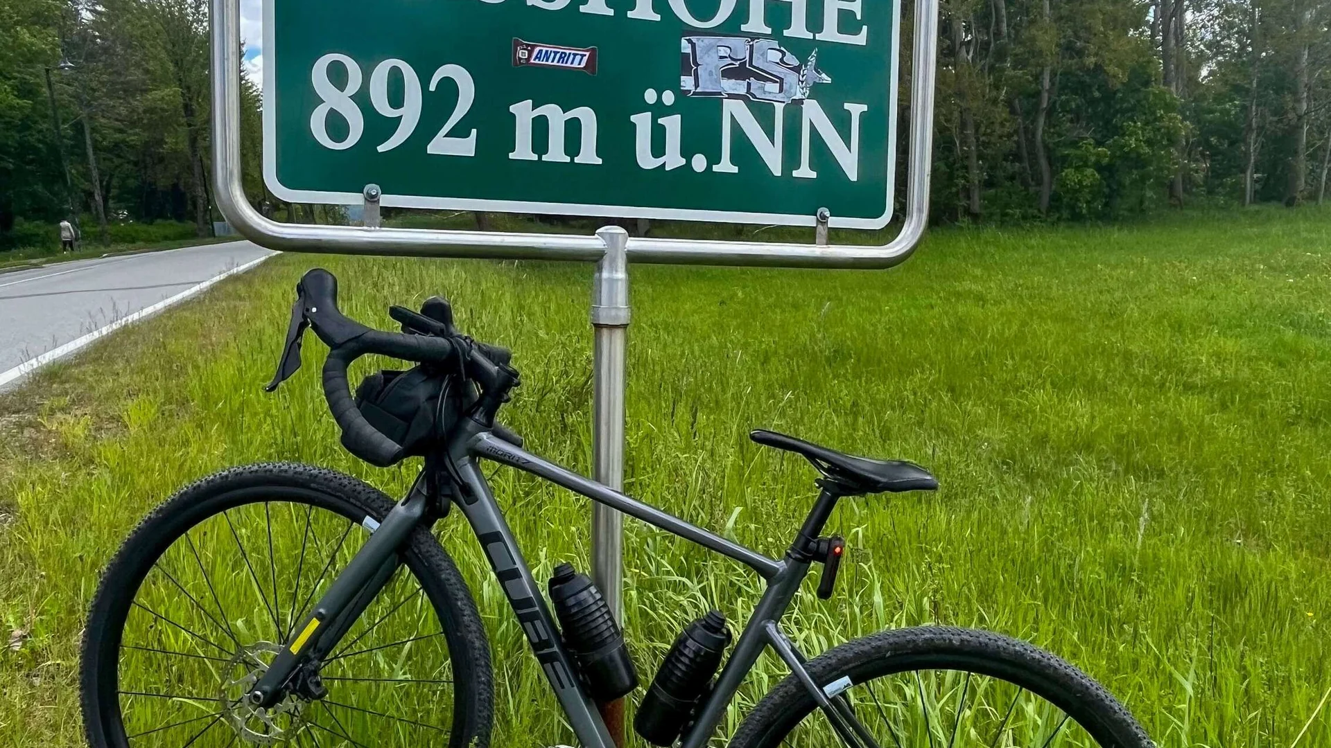 Fahrrad vor Schild mit Passhöhe 892 m ü.NN neben Straße im Gras