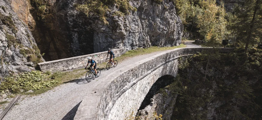 Two cyclists riding on a stone bridge in mountainous terrain