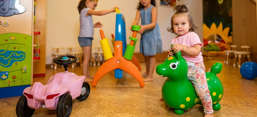 Kleinkind sitzt auf grünem Spielzeugpferd im Spielzimmer mit anderen Kindern