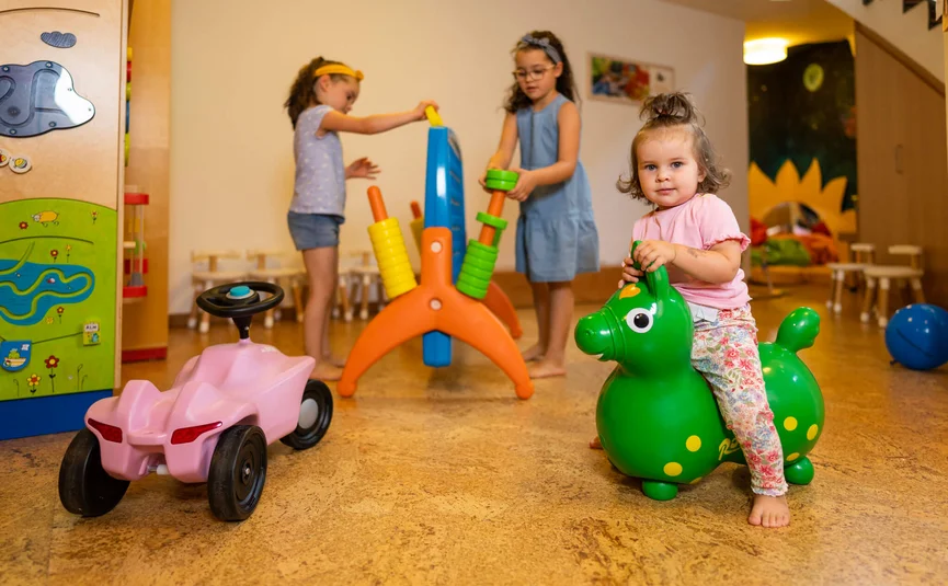 Kleinkind sitzt auf grünem Spielzeugpferd im Spielzimmer mit anderen Kindern