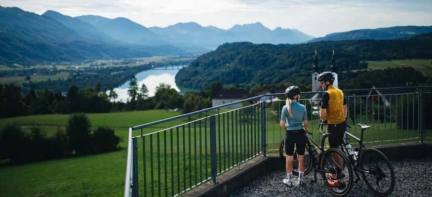 Two cyclists overlooking a landscape with river and mountains