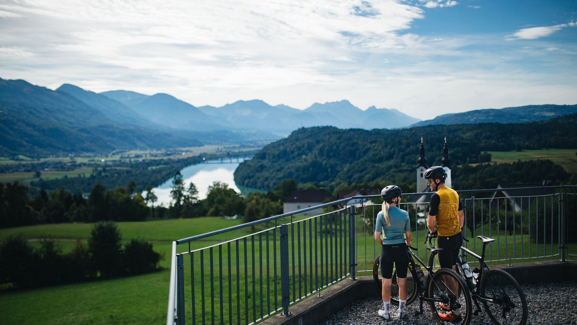 Zwei Radfahrer schauen auf eine Landschaft mit Fluss und Bergen
