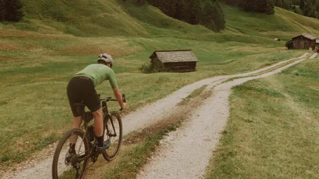 Gravelbiken ist eine Welt zwischendrin. © John Braynard Radfahrer auf Schotterweg in grüner Berglandschaft unter blauem Himmel