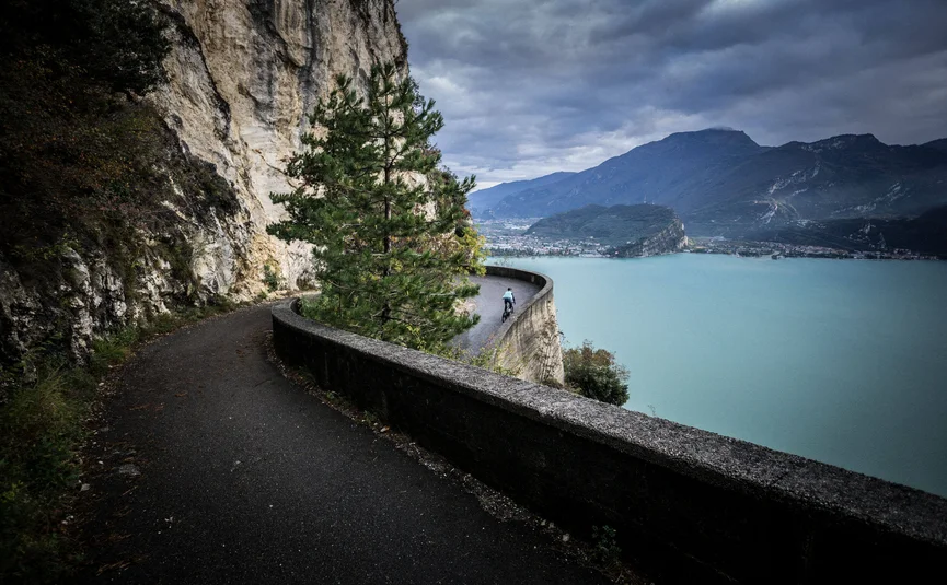 Gravel bike holiday on Northern Lake Garda © Gijs Ferkranus - Wielrenblad Magazine Cyclist on winding mountain road above a large lake with cloudy sky