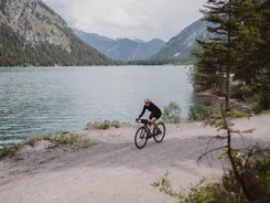 Cyclist riding on gravel path beside lake with mountains