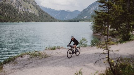 Cyclist riding on gravel path beside lake with mountains