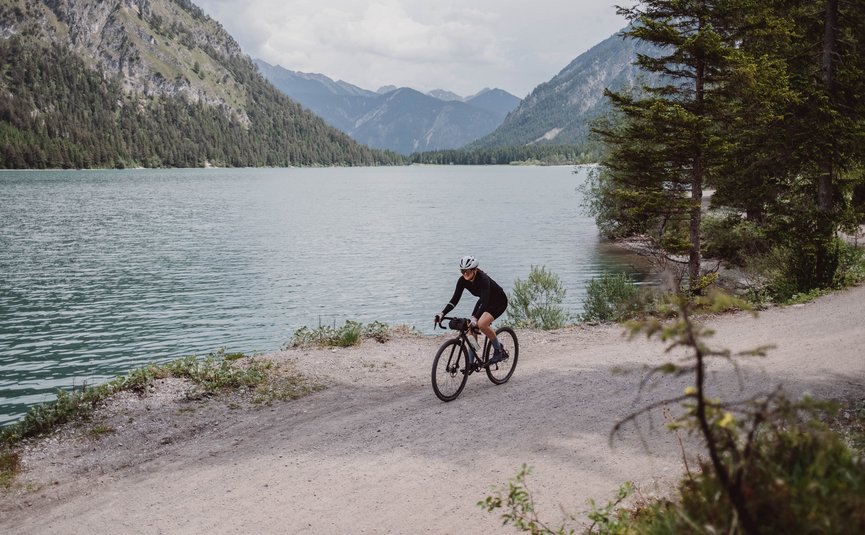 Gravelbike Urlaub in Gravel.Tirol © Liz Kellerer Fahrradfahrer auf Schotterweg neben See und Bergen