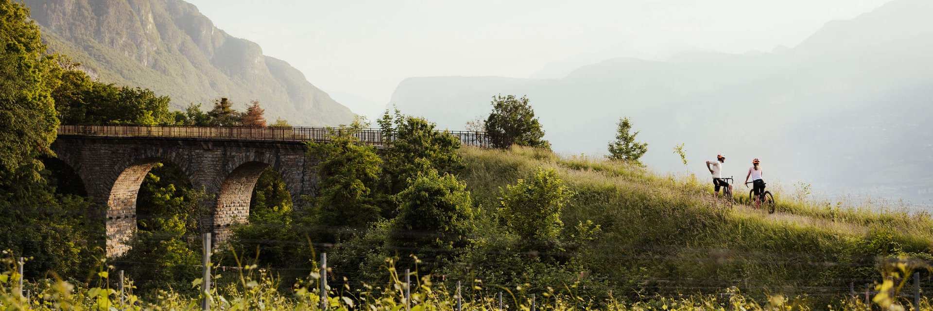 Zwei Radfahrer auf einem Hügel neben einer alten Steinbrücke in grüner Landschaft