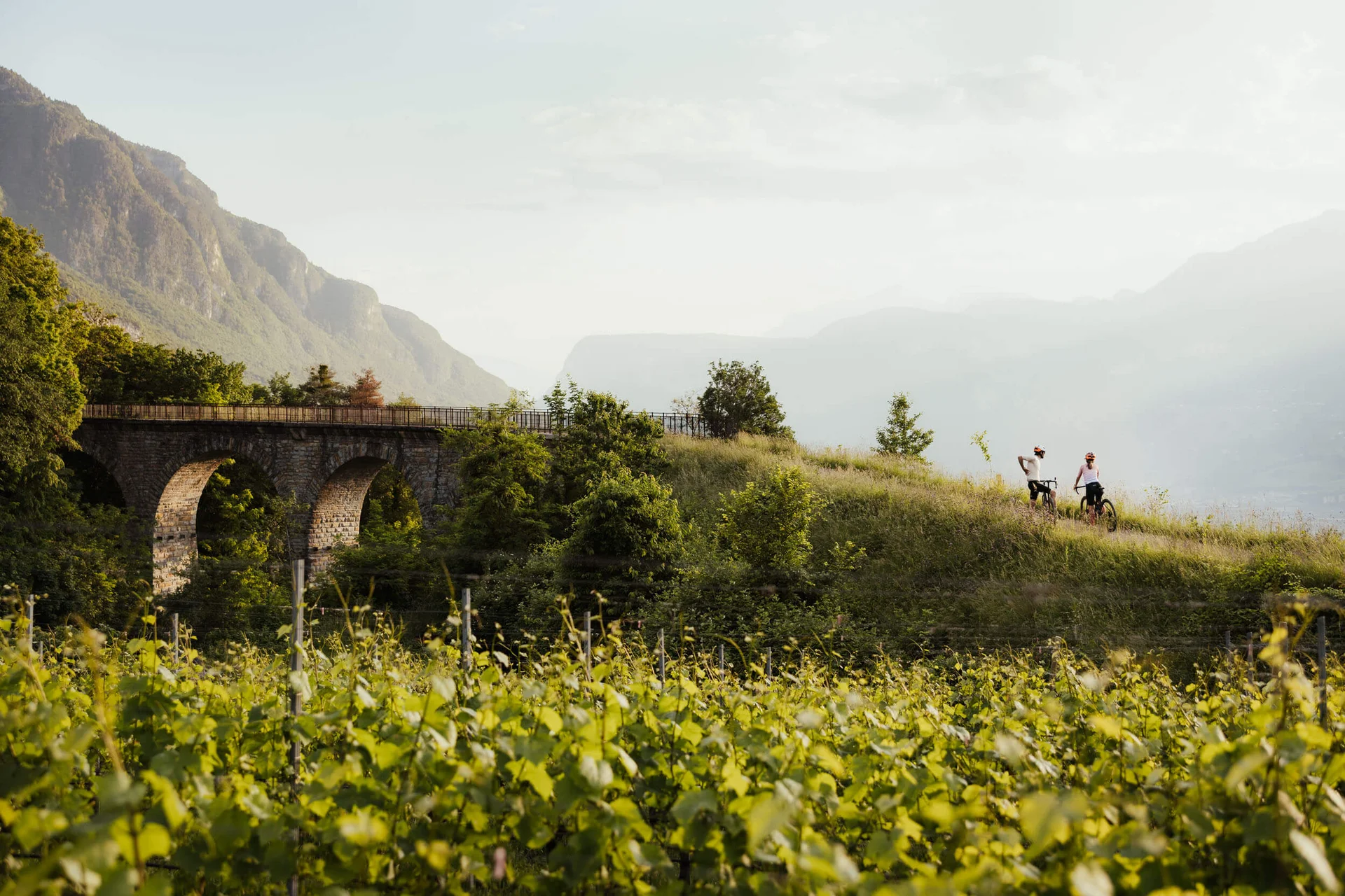 Gravelbike Holidays © Alex Moling Zwei Radfahrer auf einem Hügel neben einer alten Steinbrücke in grüner Landschaft