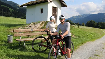 Zwei Radfahrer vor einer kleinen Kapelle auf dem Land mit Bergen im Hintergrund