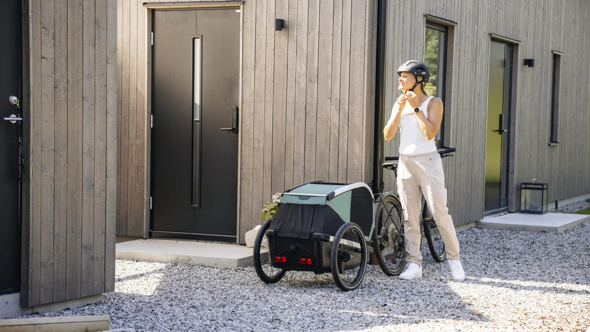 Mit dem Thule Bexey beginnt jedes Abenteuer zu zweit © Thule Frau mit Fahrradhelm neben einem Fahrrad mit Anhänger vor einem Holzgebäude