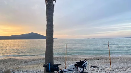 Bikepacking von Wien nach Athen © Jan Koller Fahrrad lehnt an Palme am steinigen Strand bei Sonnenuntergang