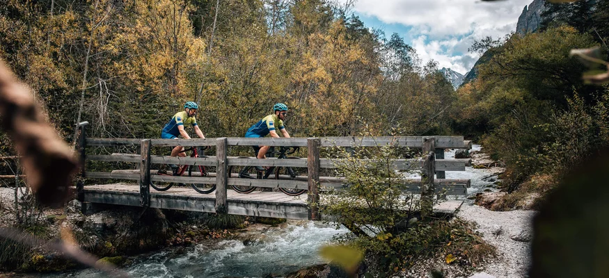 Zwei Radfahrer überqueren eine Holzbrücke über einen Bach im Wald