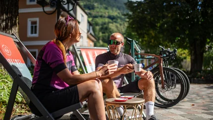 Two cyclists in sportswear drinking coffee outdoors on deck chairs.