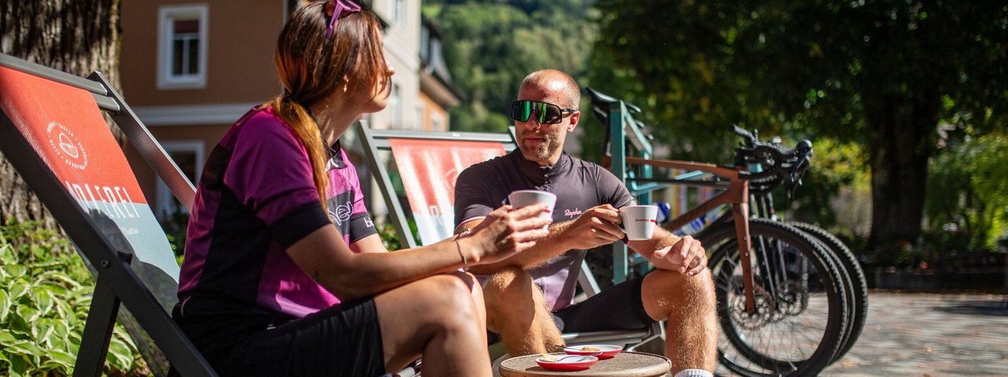 Zwei Radfahrer in Sportkleidung trinken Kaffee im Freien auf Liegestühlen.