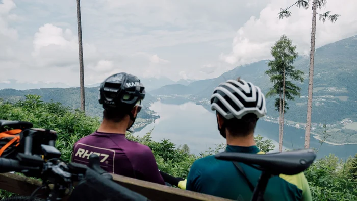 Two cyclists wearing helmets sitting on a bench overlooking a lake in the mountains